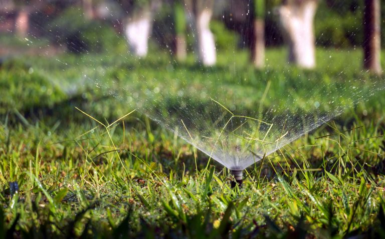 Hidden costs of a well-maintained front lawn of a suburban house is shown with sprinklers actively watering the grass. The water sprays in arching patterns, creating a shimmering effect under the sunlight.